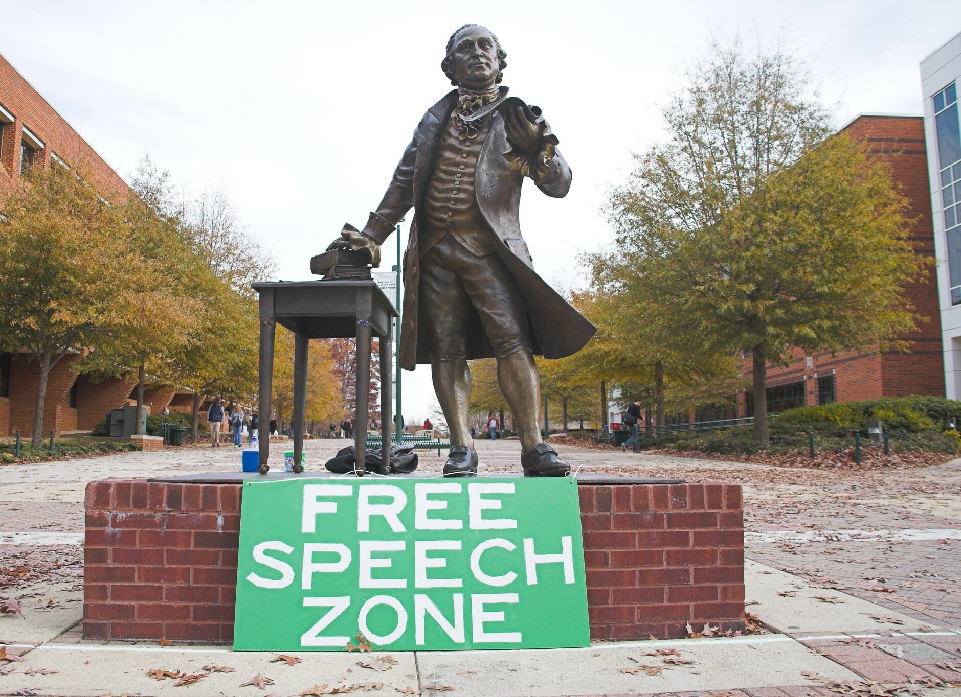 A bronze statue of a historical figure stands on a brick platform outdoors, with autumn trees in the background. A green sign in front reads FREE SPEECH ZONE in bold white letters.