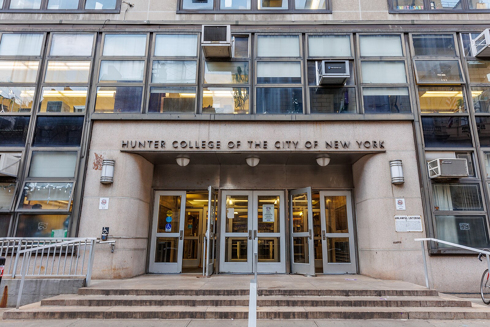 The entrance of Hunter College of the City of New York, featuring large glass windows, multiple doors, and signs above the doorway on a concrete building facade.