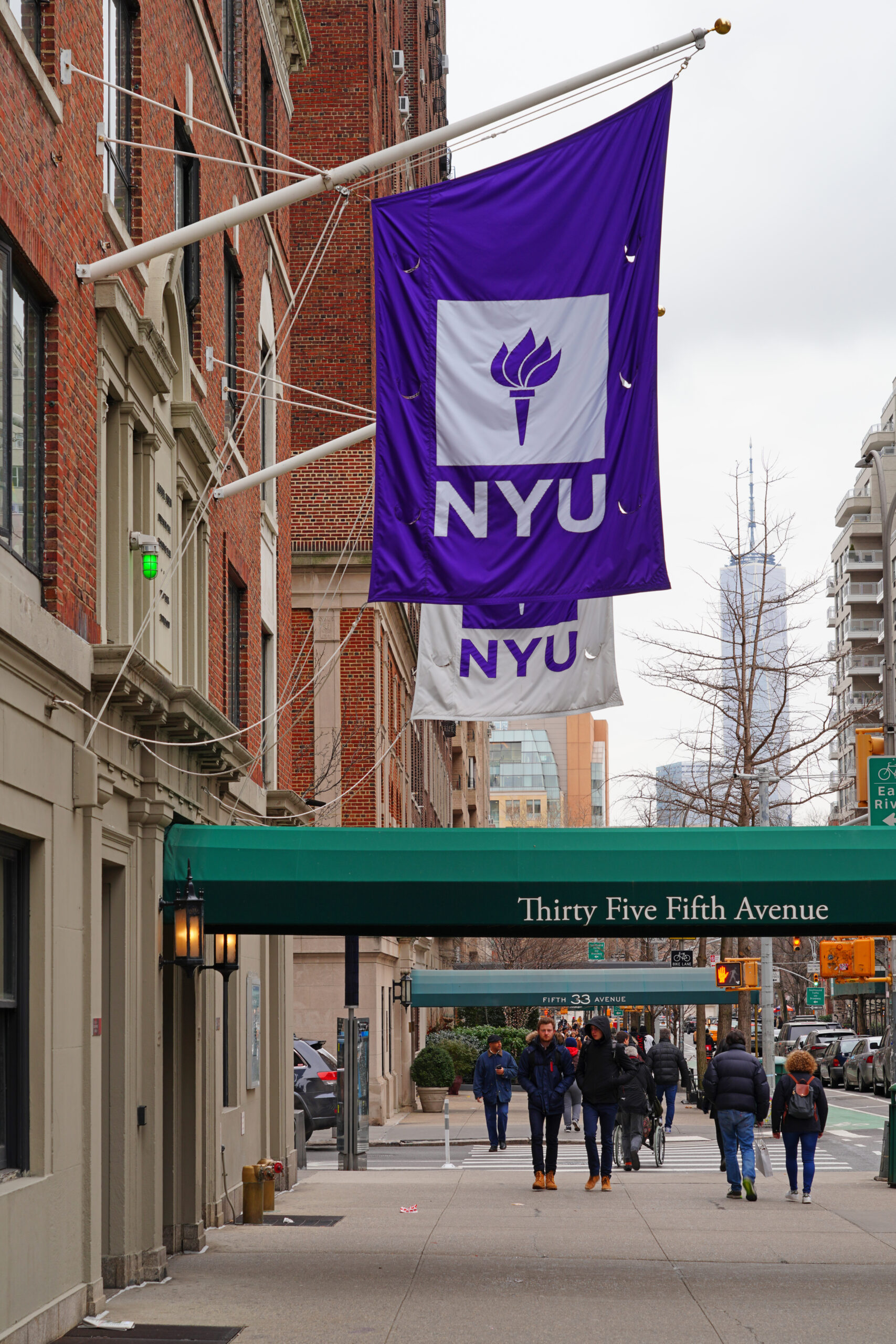 People walk on a sidewalk beneath large purple and white NYU flags outside a brick building with a green awning that reads Thirty Five Fifth Avenue in an urban city setting.
