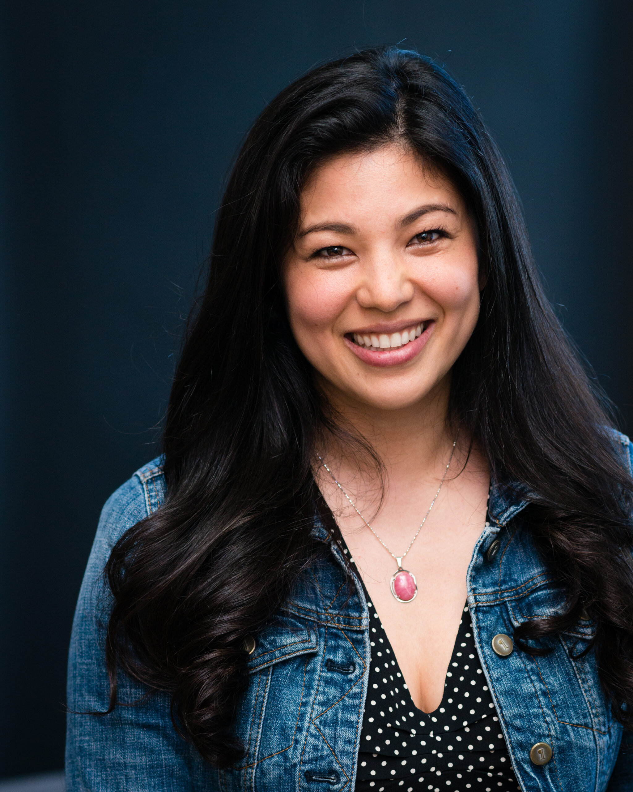 A woman with long dark hair smiles at the camera. She is wearing a denim jacket over a black blouse with white polka dots and a necklace with a pink pendant, standing against a dark, blurred background.