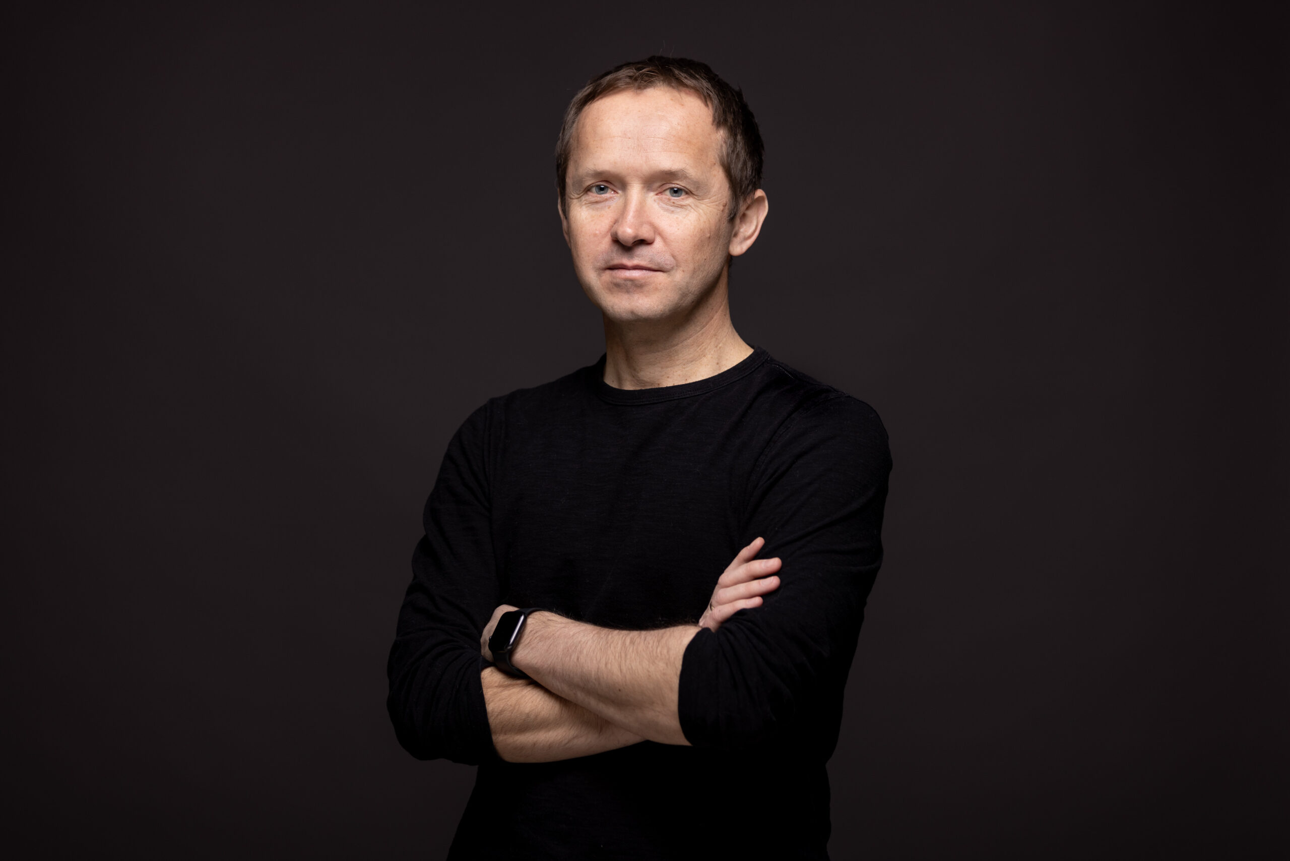 A man with short brown hair wearing a black long-sleeve shirt stands against a dark background with arms crossed, looking at the camera with a neutral expression.