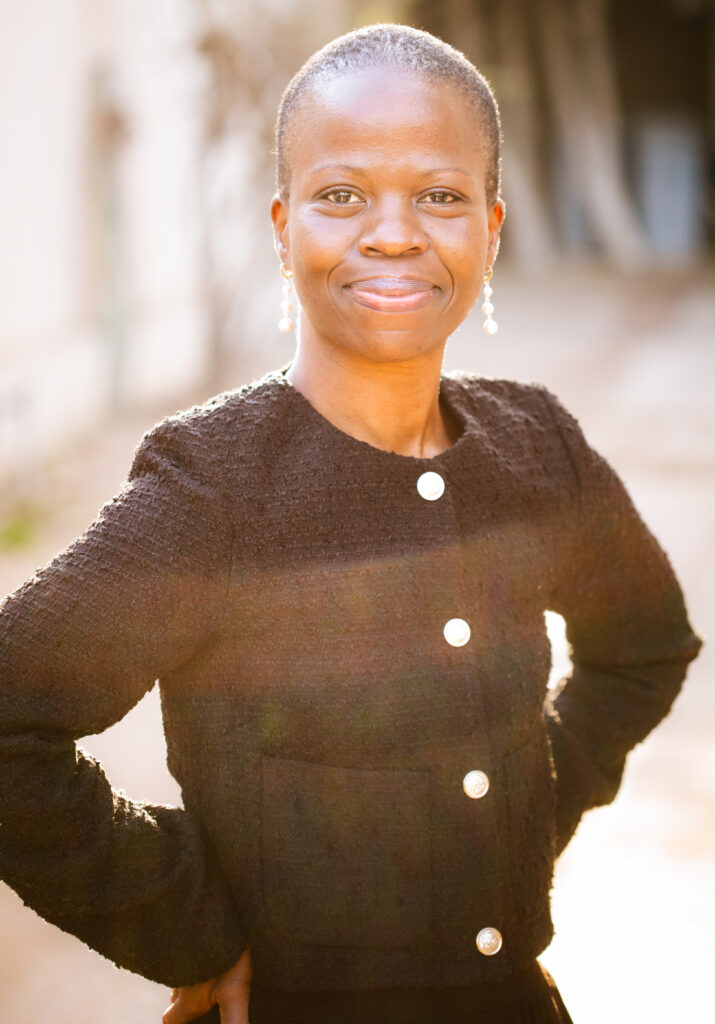 A person with short hair, wearing a textured black jacket with pearl buttons and drop earrings, stands outside smiling confidently with hands on hips in warm sunlight.