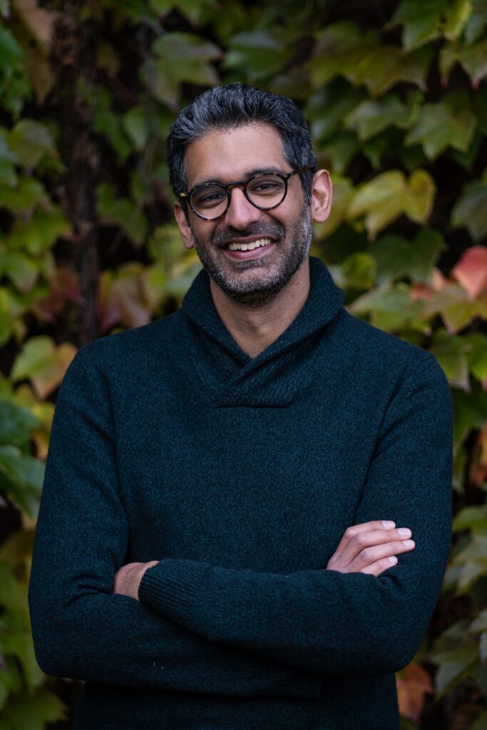 A man with short dark hair and glasses smiles with arms crossed, wearing a dark sweater, standing in front of a wall covered with green and yellow leaves.