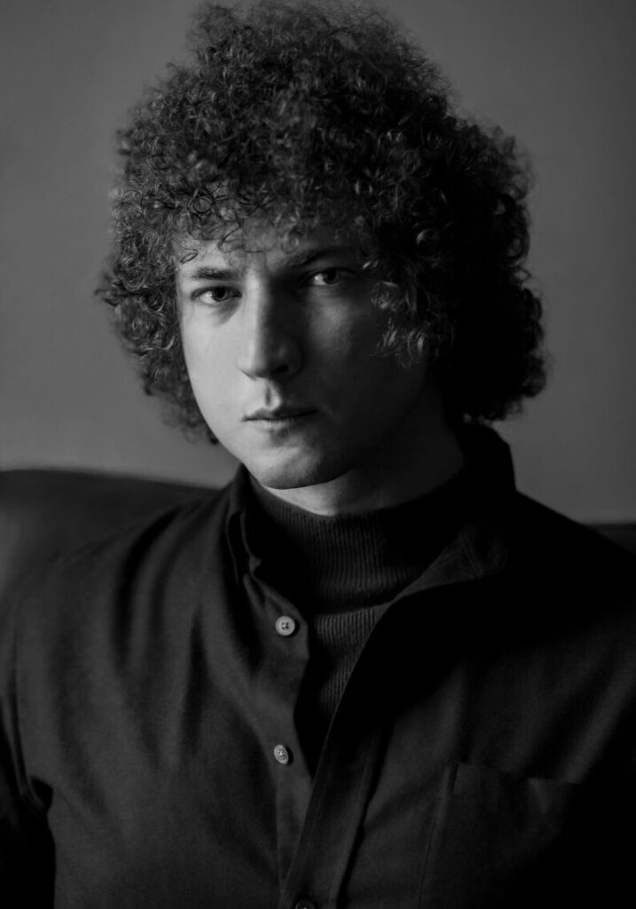 Black-and-white portrait of a young man with curly hair, wearing a dark button-up shirt and turtleneck, looking directly at the camera with a serious expression.