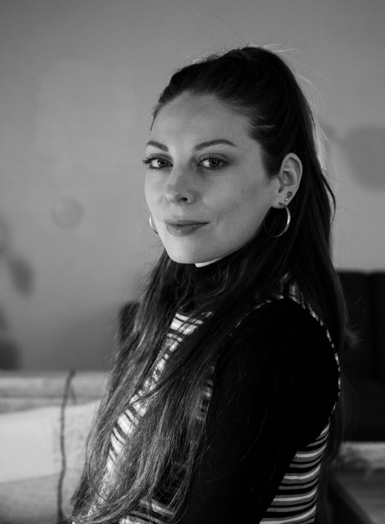 A woman with long dark hair in a ponytail, wearing hoop earrings and a striped top, looks at the camera and smiles softly. The photo is in black and white with a blurred background.