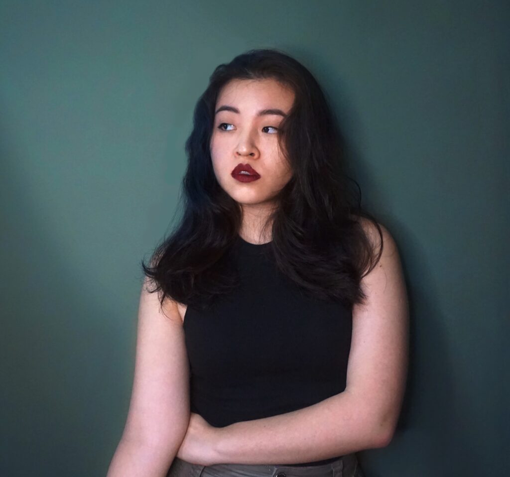 A young woman with long dark hair and dark lipstick stands against a plain green wall, wearing a sleeveless black top and looking to the side with a thoughtful expression.