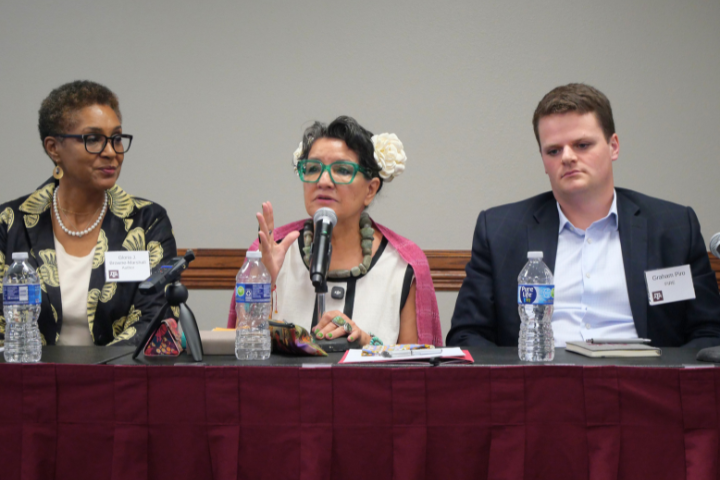 Three people sit at a conference panel table with microphones and water bottles. The woman in the center is speaking, flanked by a woman on the left and a man on the right. All three wear name tags and business attire.