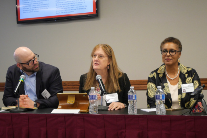 Three people sit at a table with microphones and water bottles, participating in a panel discussion. A screen with text is visible behind them. The middle person is speaking while the others listen.