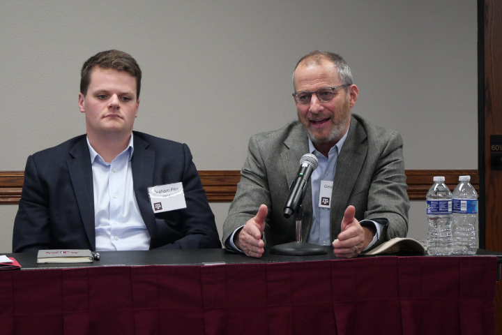 Two men in business attire sit at a table with name tags. One speaks into a microphone with hands gesturing, while the other sits quietly. Water bottles and notebooks are on the table in front of them.