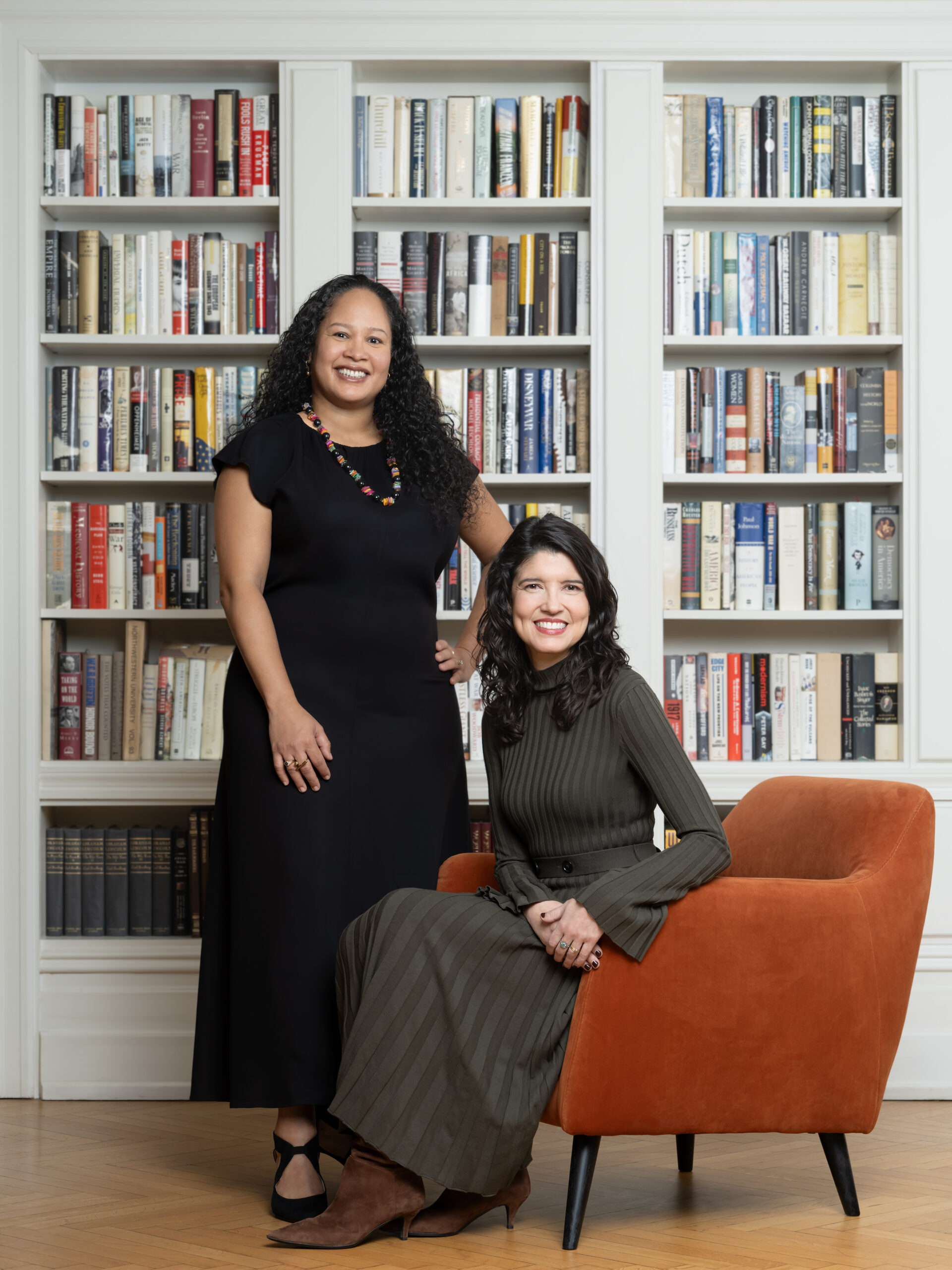 Two women pose in a library. One stands, wearing a black dress and colorful necklace, with long curly hair. The other sits in an orange chair, wearing a green dress and brown boots. Bookshelves filled with books are behind them.