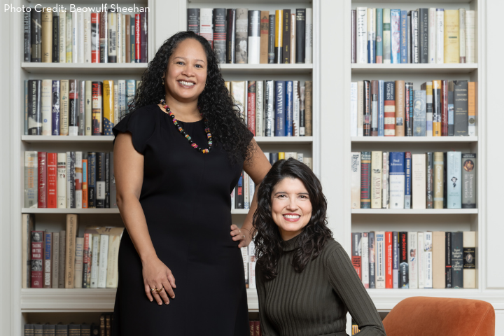 Two women pose in front of bookshelves filled with books. One woman stands, wearing a black dress and necklace, while the other sits, wearing a dark green ribbed top. Both are smiling.