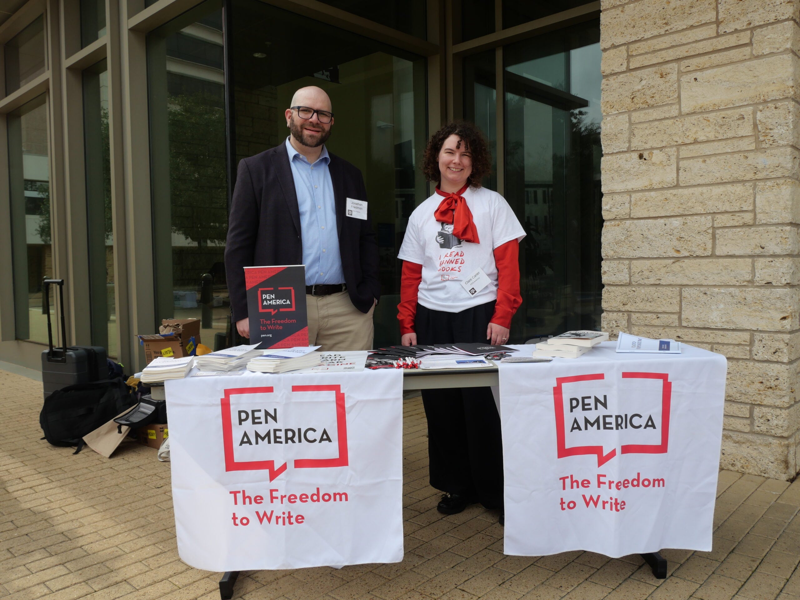 Two people stand behind a table covered with PEN America banners and materials, promoting “The Freedom to Write.” The table is set up outdoors near a building with large windows.