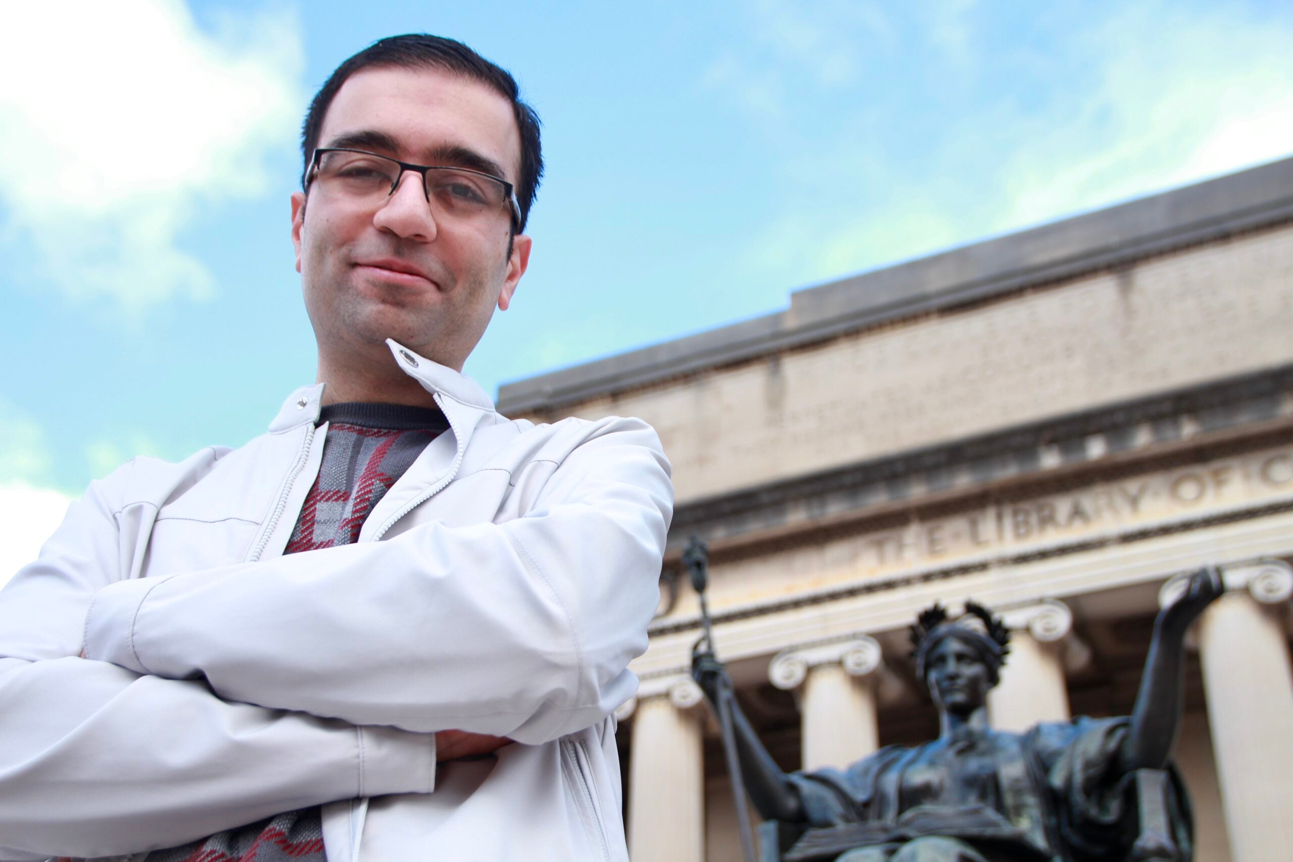 A man with glasses and dark hair, wearing a white jacket, stands with arms crossed in front of a classical building and a statue holding a staff. The sky is blue with clouds.