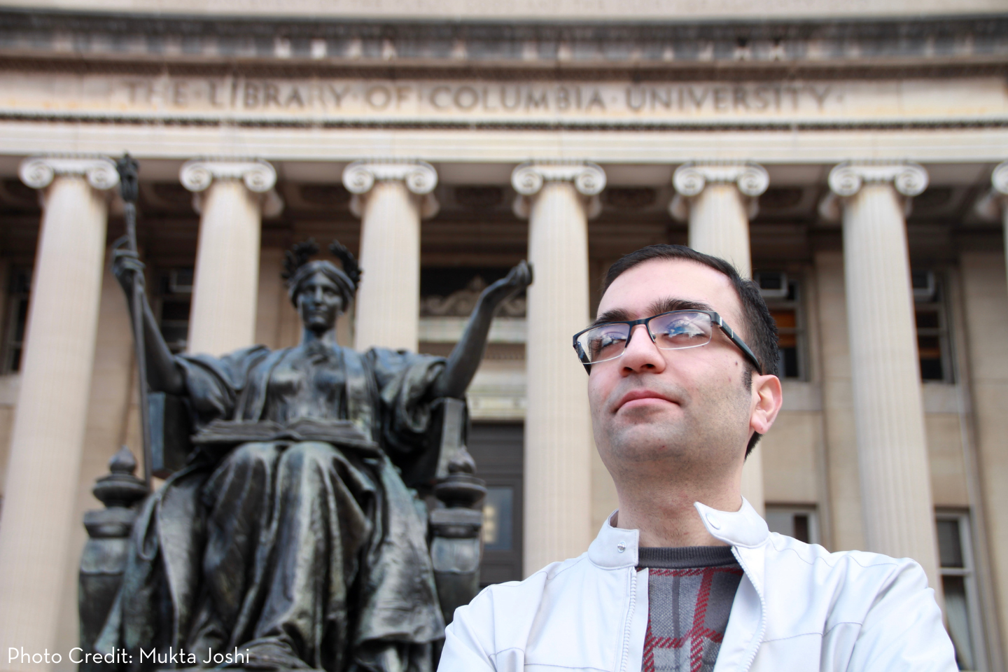 A man in glasses and a white jacket stands in front of the Alma Mater statue and the columns of Columbia University’s library, looking slightly upward with a thoughtful expression.