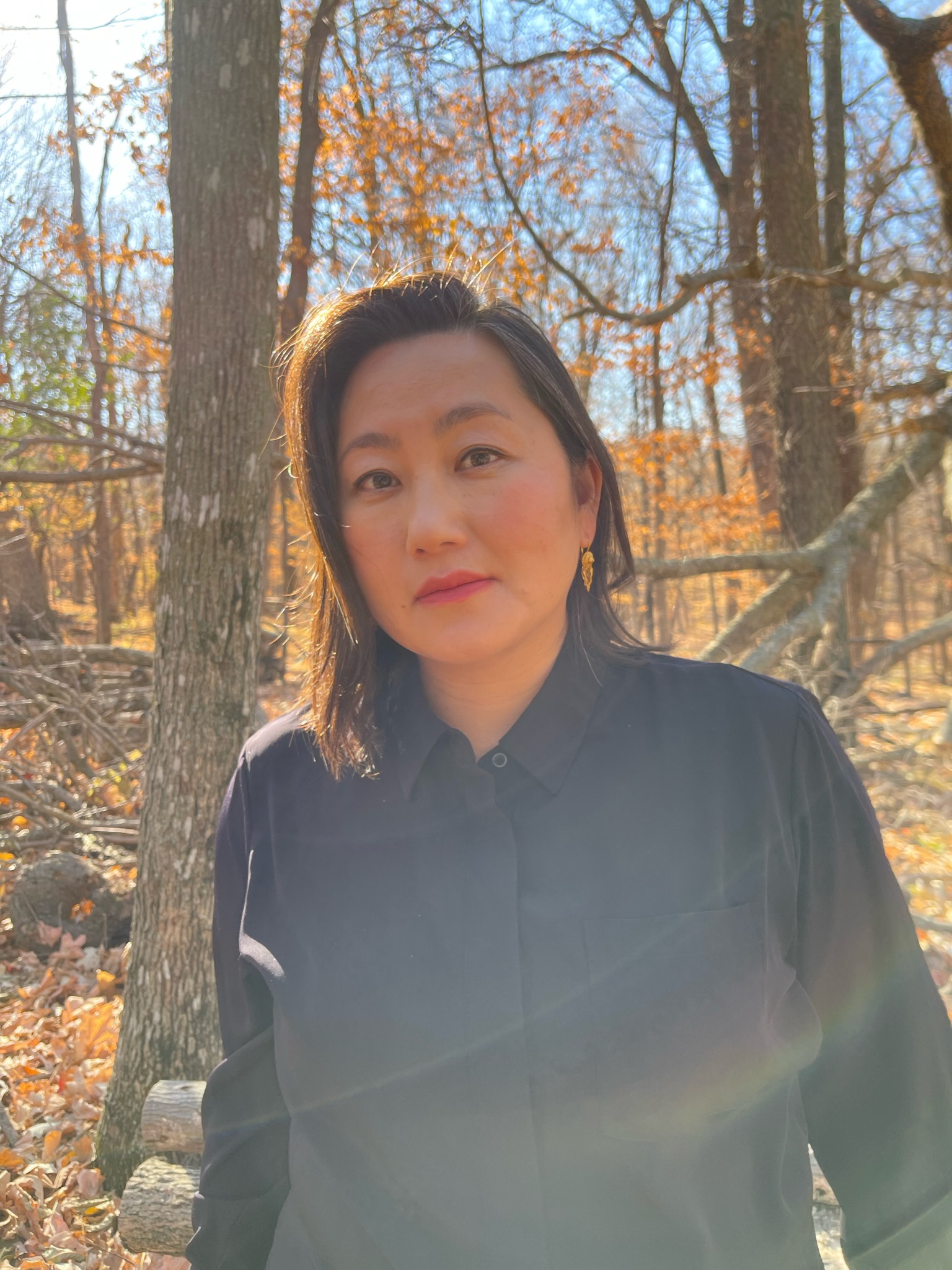 A person with straight, shoulder-length dark hair and wearing a black button-up shirt stands in a sunlit autumn forest with bare trees and fallen leaves. Sunlight creates a soft lens flare across the image.