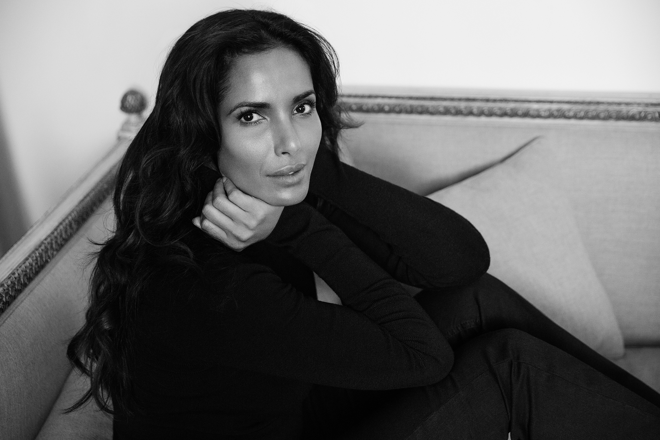 A woman with long dark hair sits on a couch, dressed in dark clothes, looking directly at the camera with a calm expression. One hand rests on her neck, and the image is in black and white.