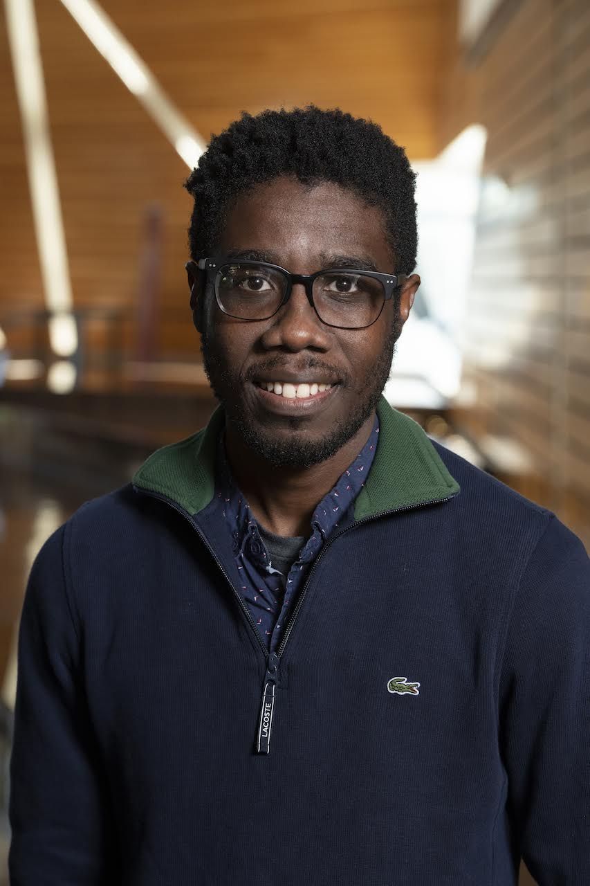 A man with short curly hair and glasses smiles, wearing a navy blue zip-up with a green collar and a patterned shirt underneath, standing indoors in front of a blurred wooden background.