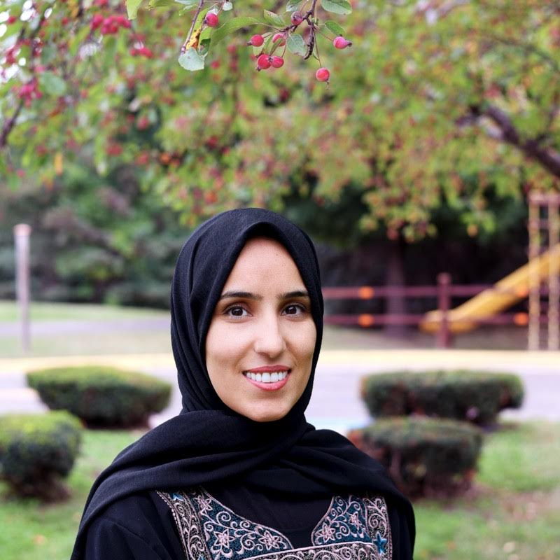A woman wearing a black hijab and an embroidered top smiles outdoors in a park, with green trees, trimmed bushes, and red berries visible in the background.