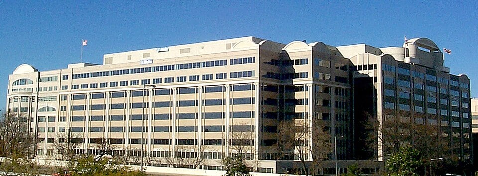A large, modern office building with many windows and multiple stories, featuring flags on the roof, photographed on a clear, sunny day with a blue sky in the background.