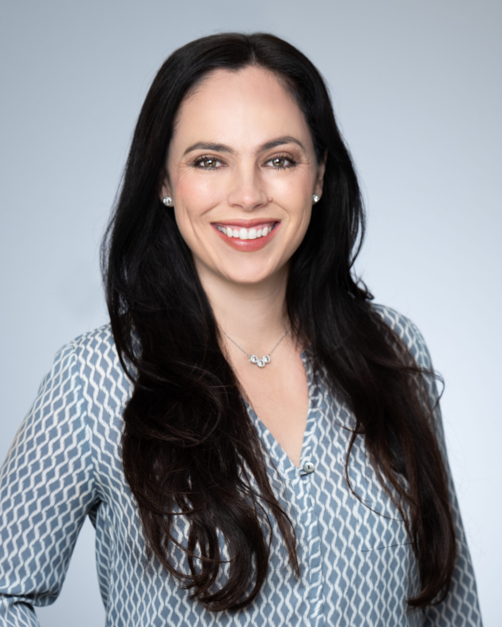 A woman with long dark hair wearing a patterned blue blouse and a necklace, smiling at the camera against a plain light gray background.