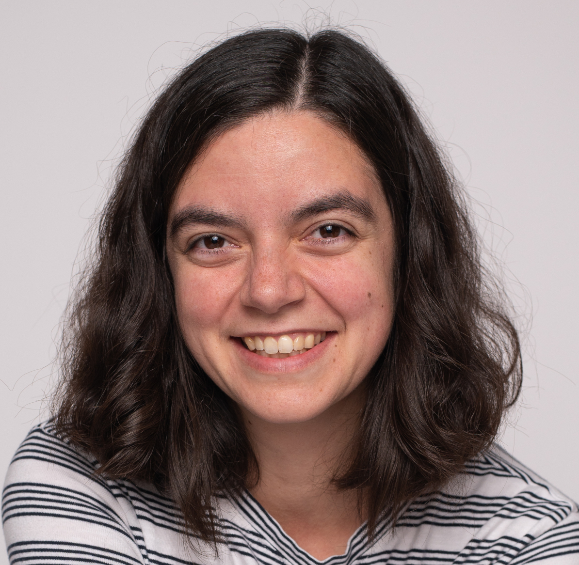 A woman with shoulder-length dark wavy hair, wearing a white shirt with black stripes, smiles warmly at the camera against a plain light background.