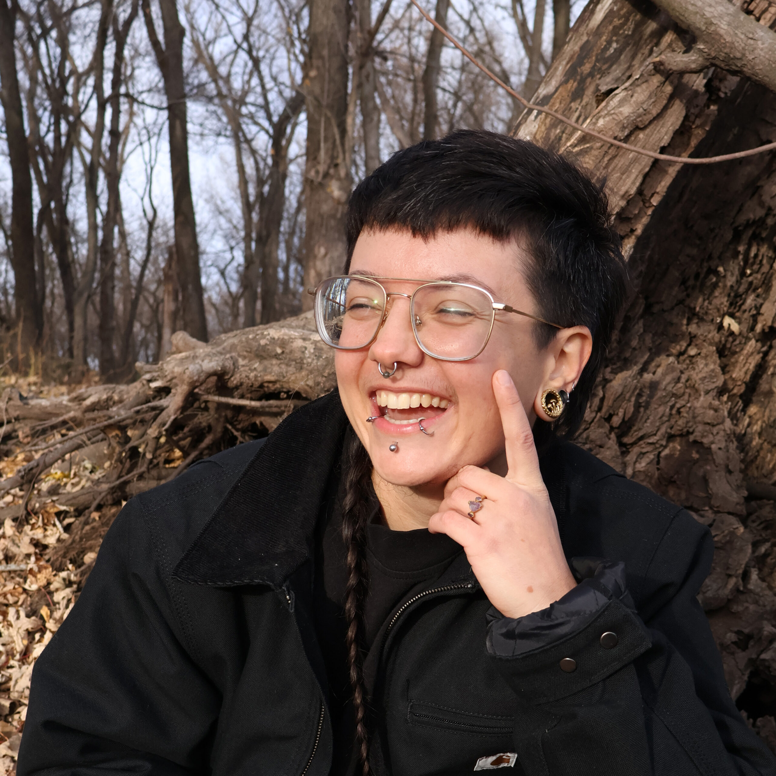 A person with short dark hair, glasses, facial piercings, and a braided strand of hair smiles while sitting outdoors against a tree in a forest with bare branches.