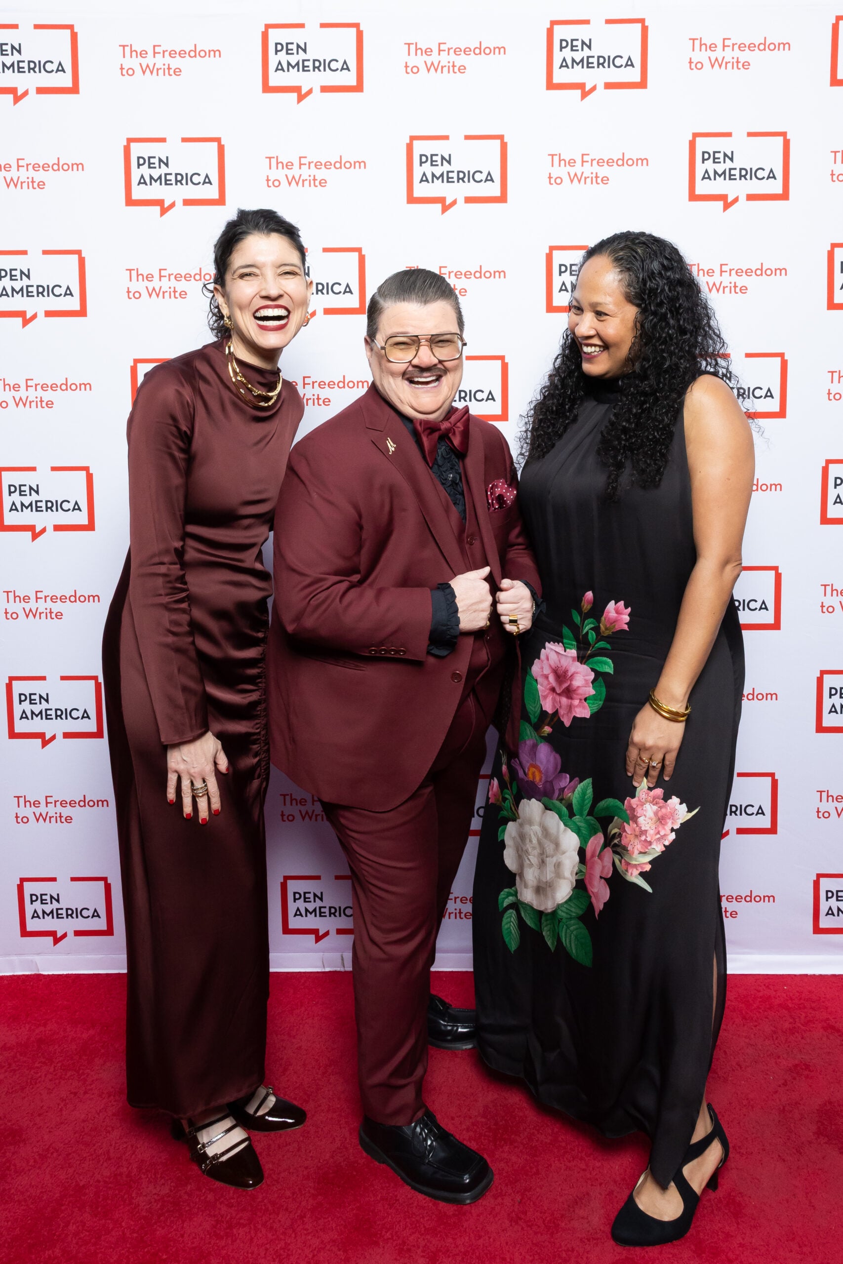 Three people stand smiling on a red carpet in front of a PEN America backdrop. The person in the center wears a burgundy suit, while the others wear long brown and black dresses, one with floral patterns.