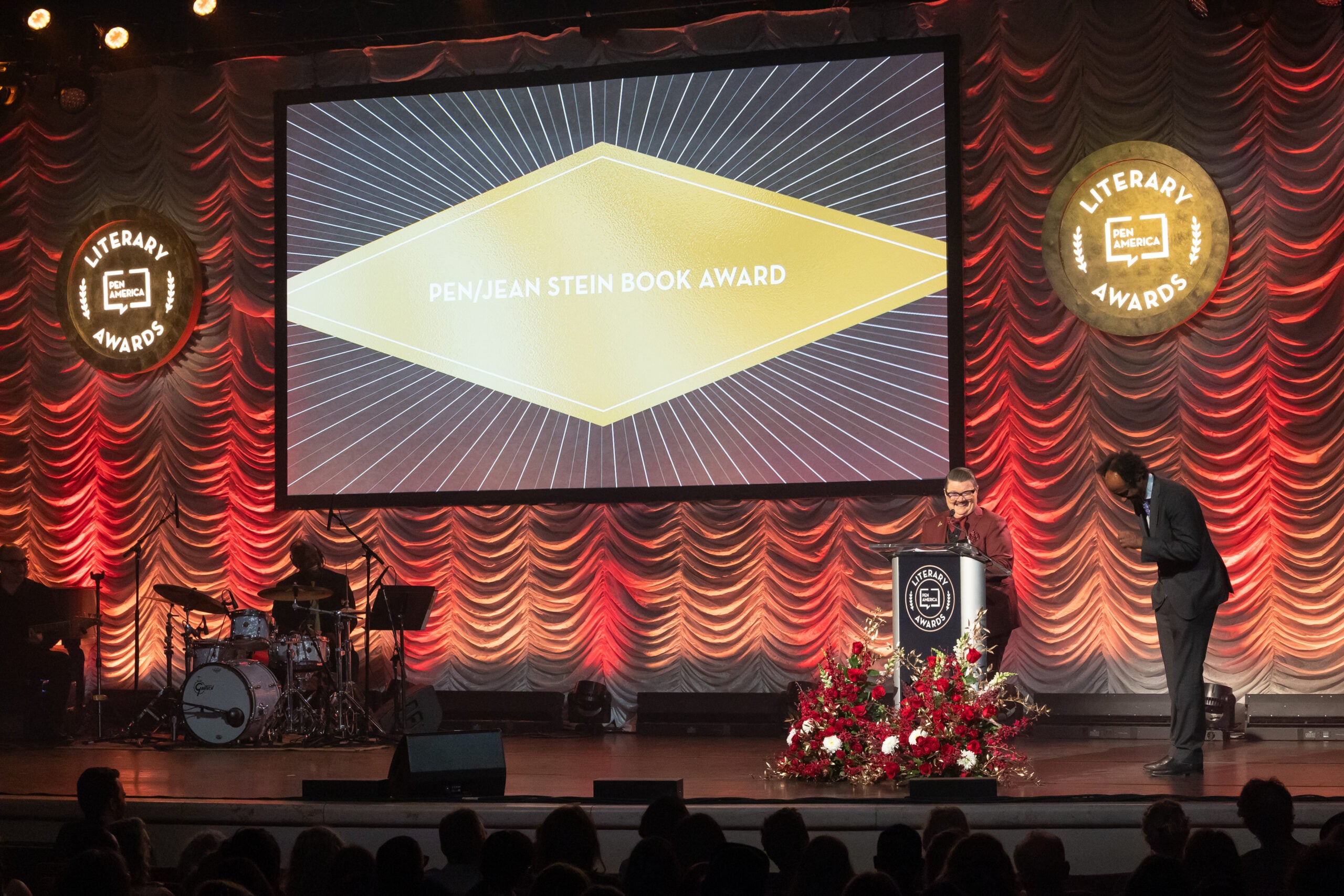 A person stands at a podium announcing the PEN/Jean Stein Book Award on a stage with red drapes, floral arrangements, and two large PEN America Literary Awards signs, while a band plays in the background.