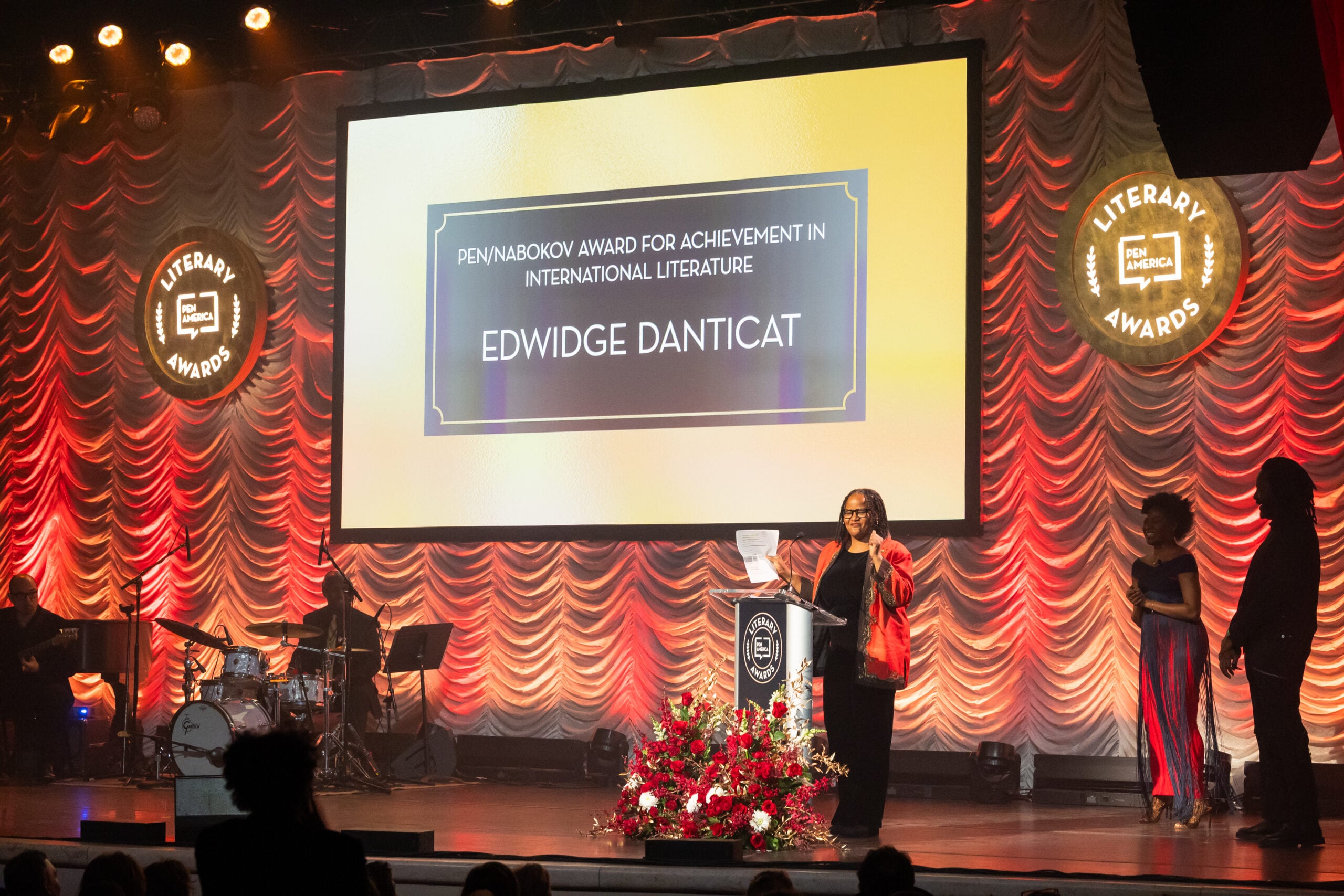 A woman stands at a podium onstage holding a paper, with a flower arrangement in front, as a screen behind her displays “PEN/Nabokov Award for Achievement in International Literature—Edwidge Danticat.” Musicians and others are onstage.