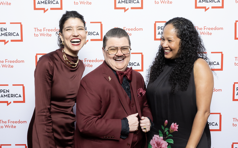 Three people stand smiling and laughing in front of a PEN America backdrop. Two wear burgundy outfits, and one wears a black dress with flower details. The backdrop reads PEN America and The Freedom to Write.