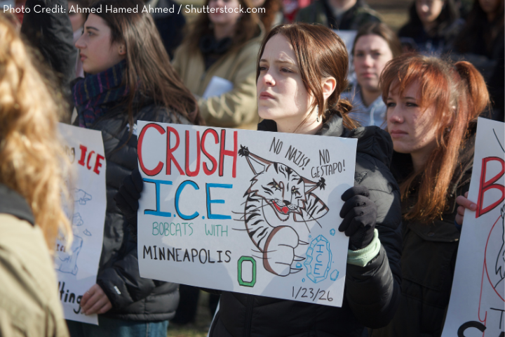 A group of protestors holds handmade signs, including one reading “CRUSH ICE, Bobcats with Minneapolis,” featuring a drawing of a bobcat and anti-ICE messages. The crowd appears serious and focused.
