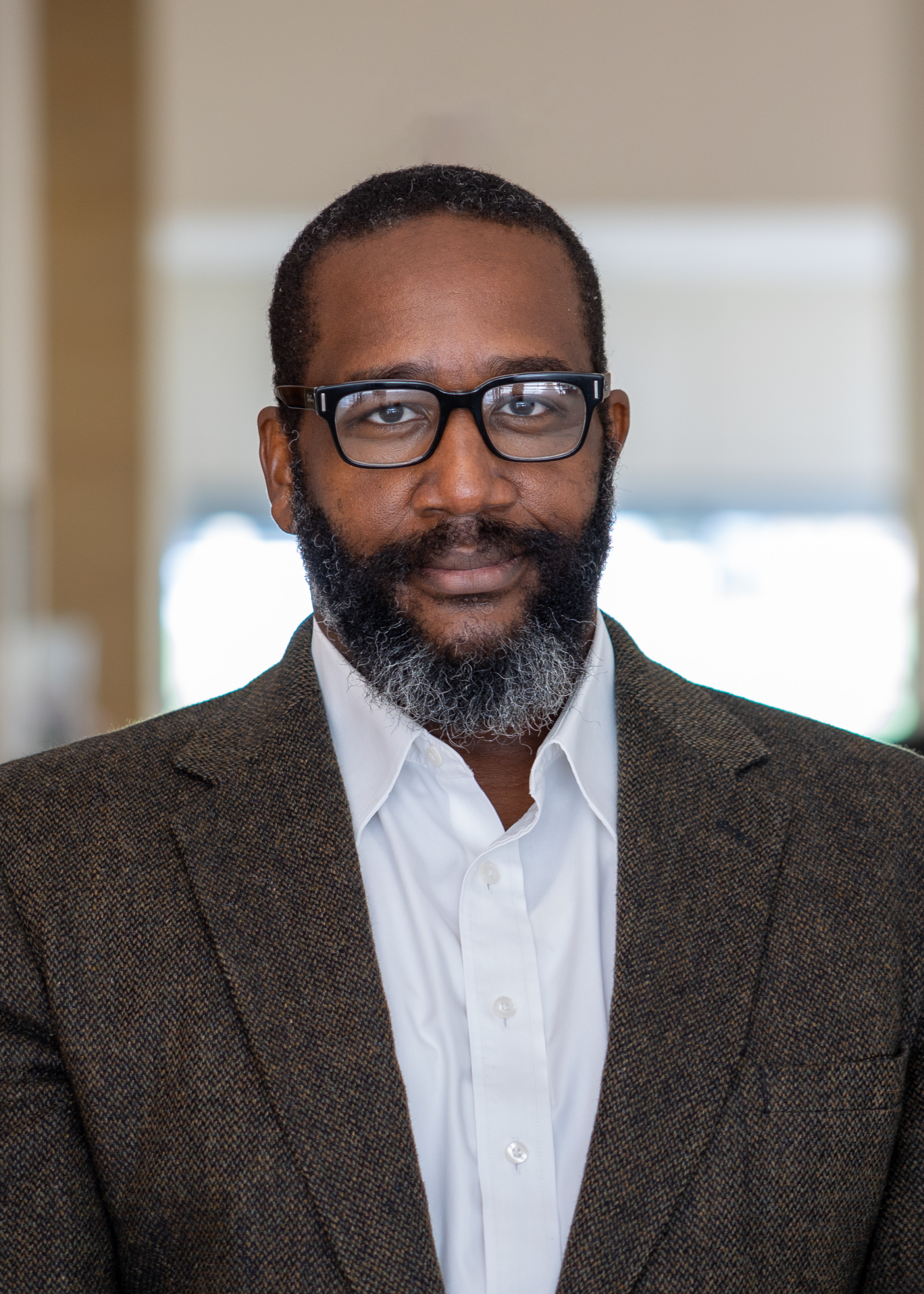 A man with short hair, a full beard, and glasses is wearing a brown textured blazer over a white dress shirt, standing indoors with a softly blurred background.
