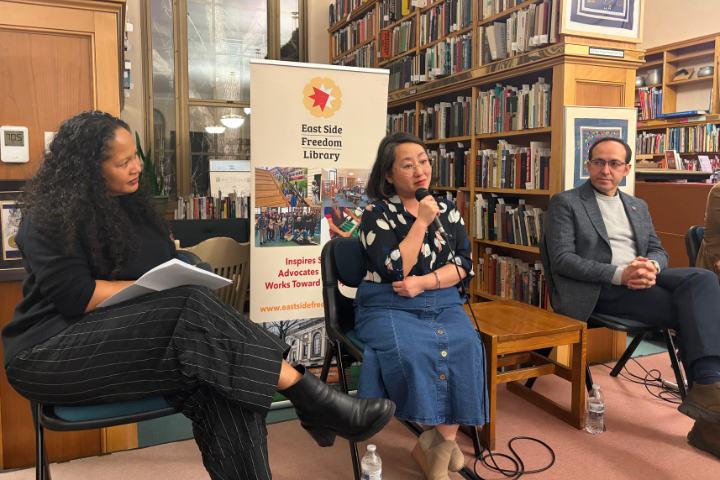 Three people sit in chairs having a panel discussion in a library. Bookshelves fill the background and a banner reads “East Side Freedom Library.” A woman in the middle speaks into a microphone.