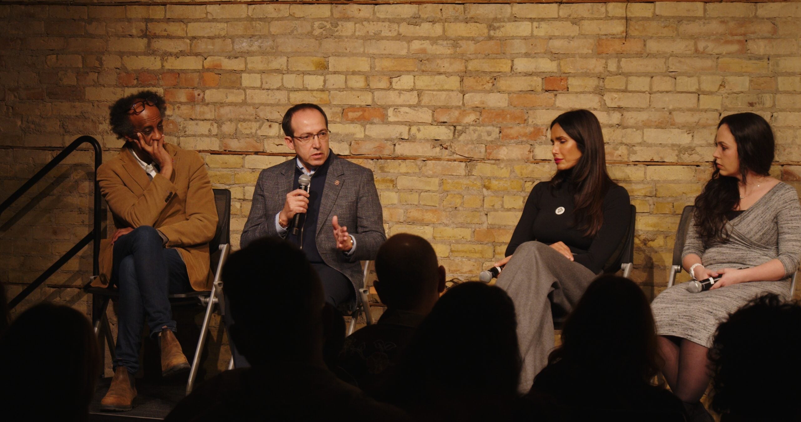 Four people sit on a panel in front of an audience, with one man holding a microphone and speaking. The other three panelists, two women and one man, listen attentively. The background is an exposed brick wall.
