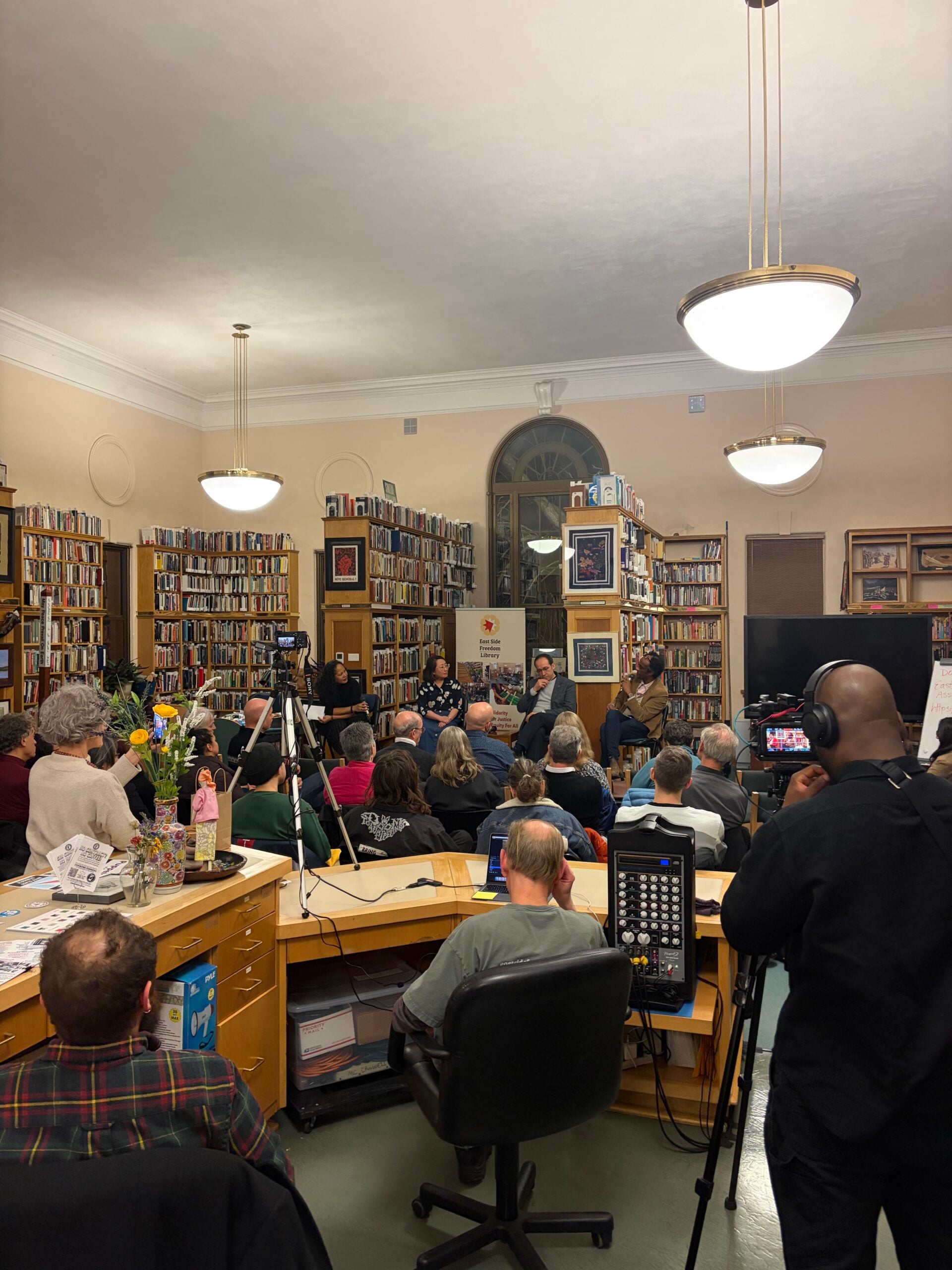 A group of people attend a panel discussion or reading in a cozy, well-lit bookstore with bookshelves lining the walls. Cameras and microphones are set up to record the event, and an audience listens attentively.
