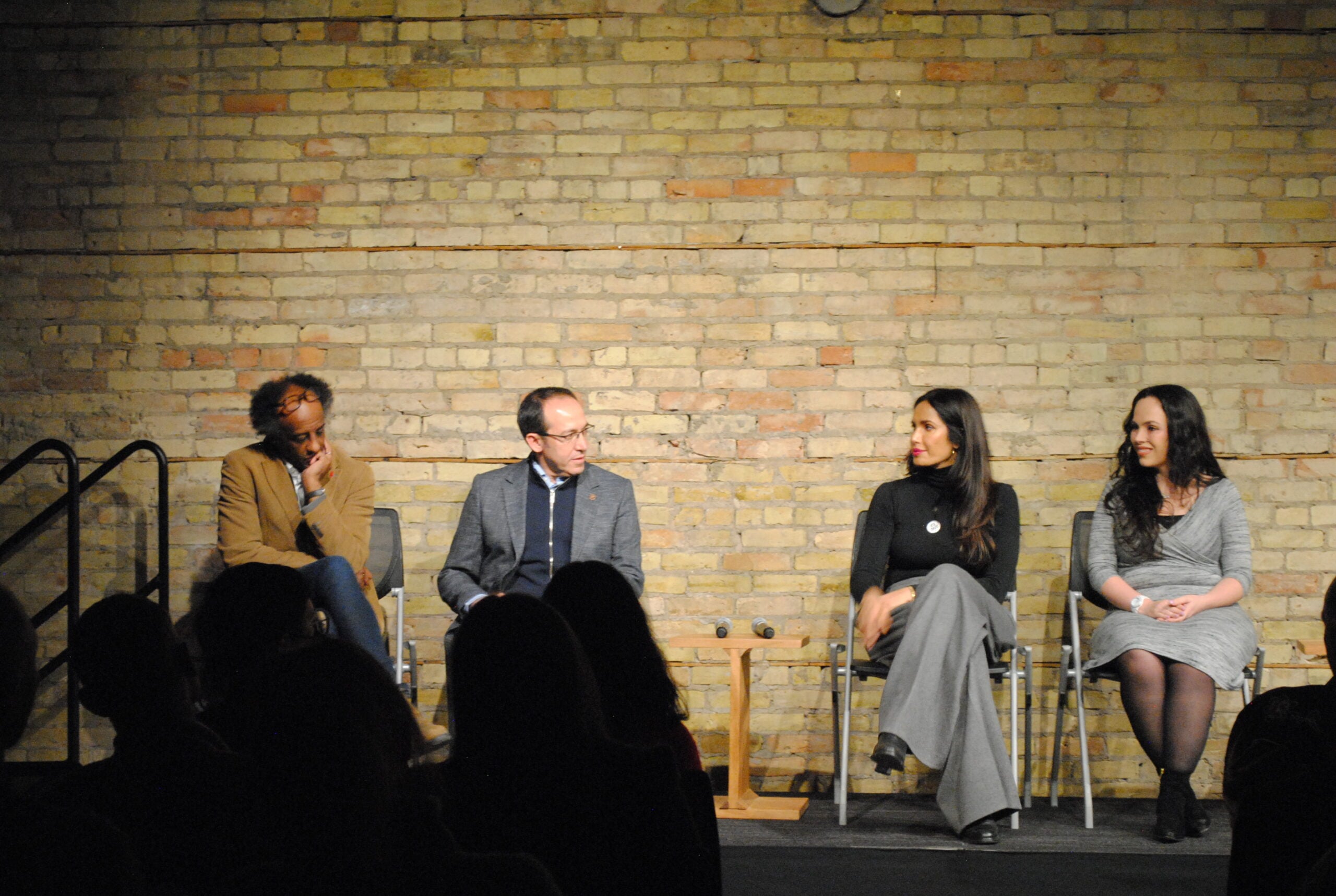 Four people sit on chairs in front of a brick wall, engaged in a panel discussion. Three appear to be listening to the fourth, who is talking. The audience is visible as silhouettes in the foreground.
