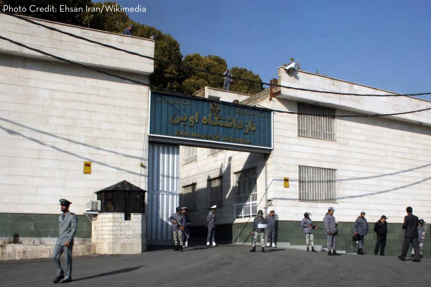 A white-walled prison building with a blue sign reading Evin House of Detention above the entrance gate. Several guards in uniform stand outside, and two guards are seen atop the wall. Trees are visible in the background.