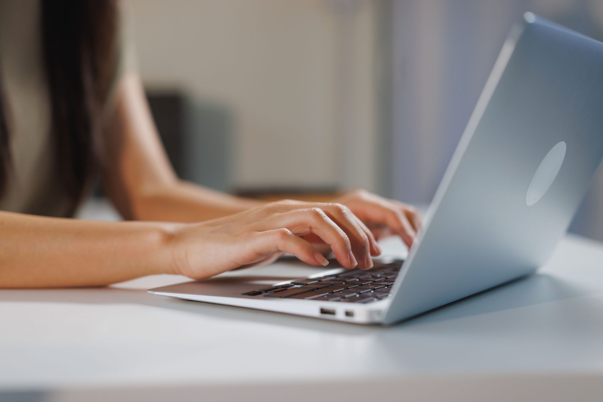 A person typing on a laptop at a white desk, with only their hands and arms visible. The background is softly blurred, focusing attention on the act of typing.