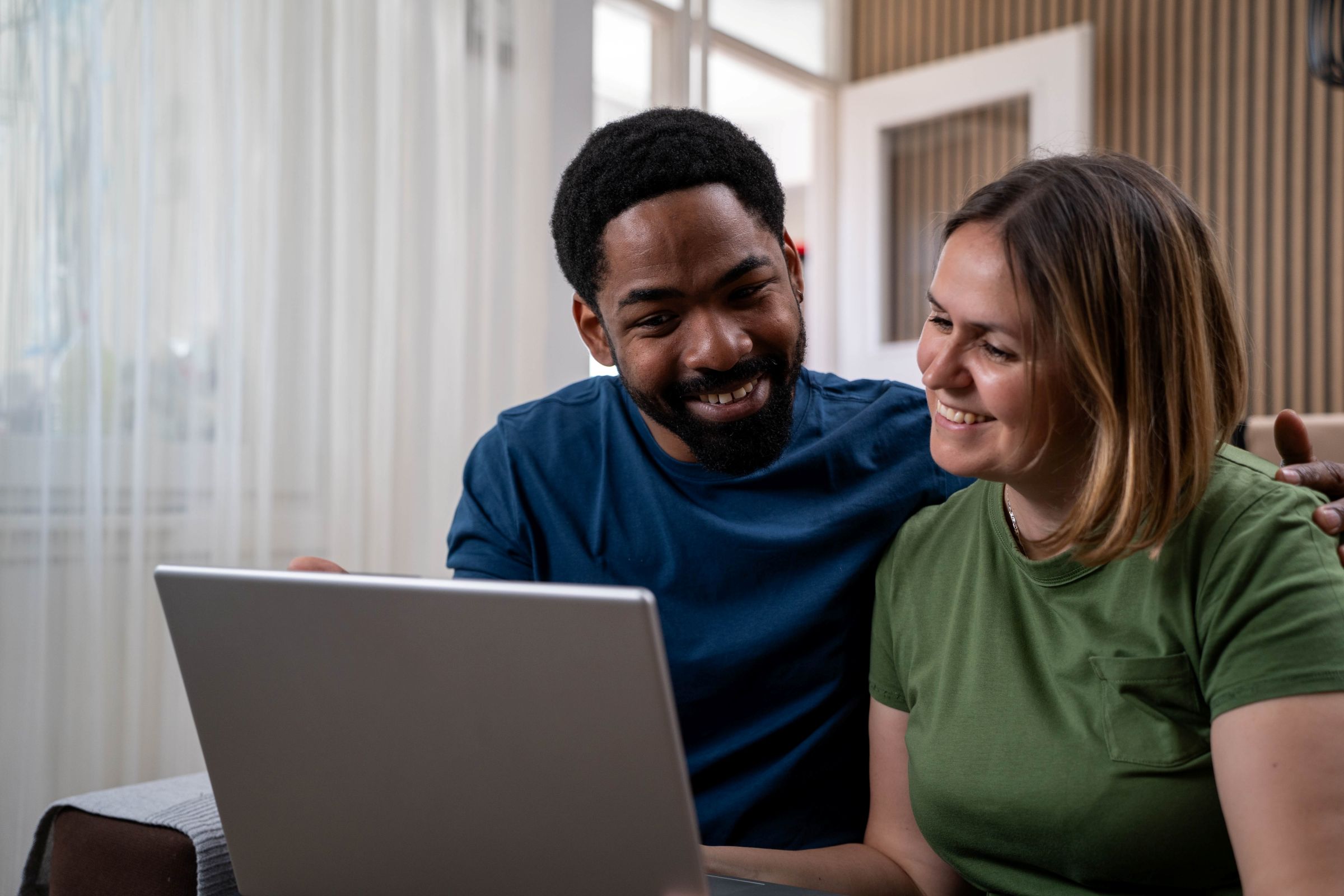 A smiling couple sits close together on a couch, looking at a laptop screen. The man has his arm around the woman, and they both appear happy and engaged with what they are viewing on the computer.
