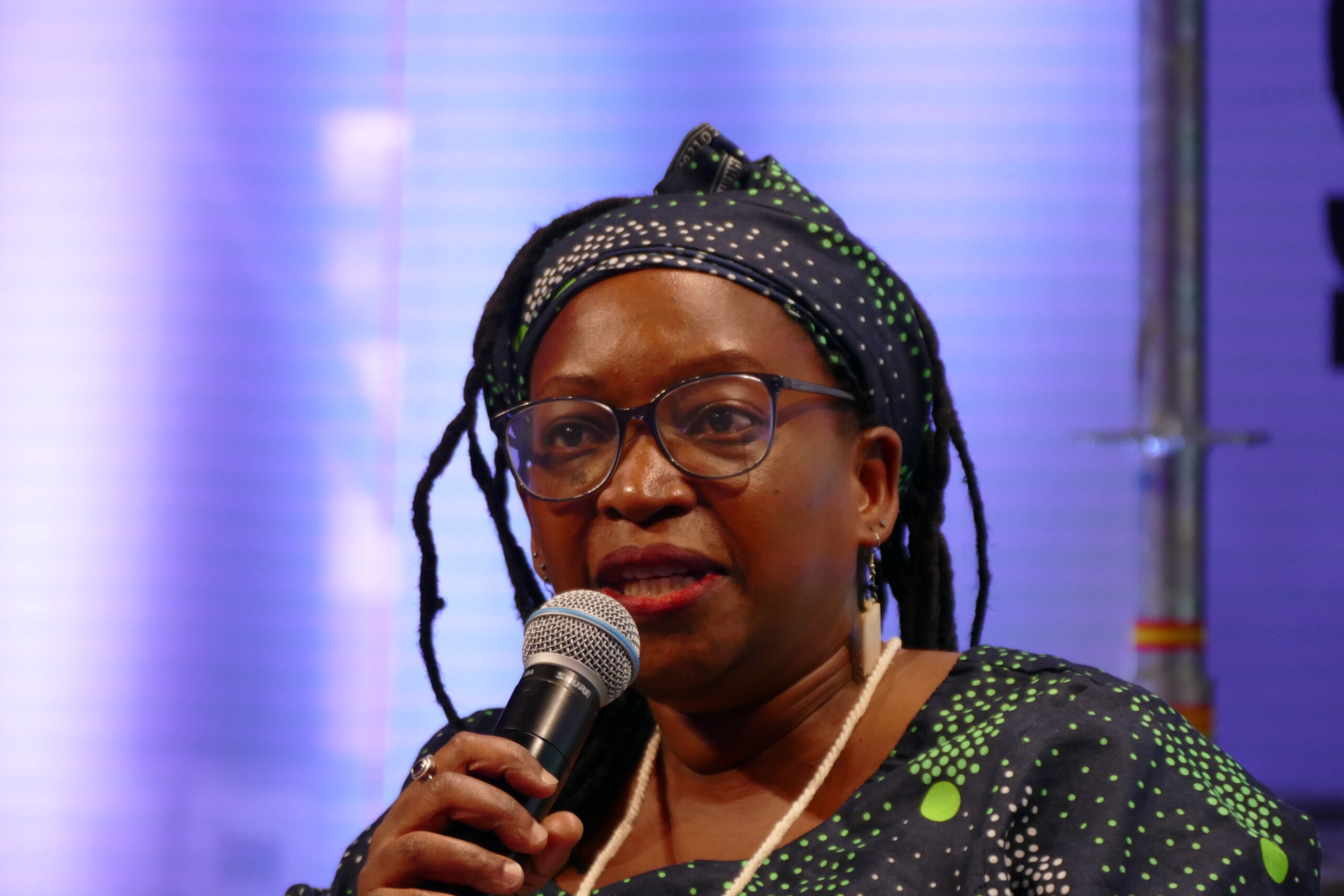 A woman wearing glasses, a patterned headscarf, and matching attire speaks into a microphone against a blurred, purple-lit background.