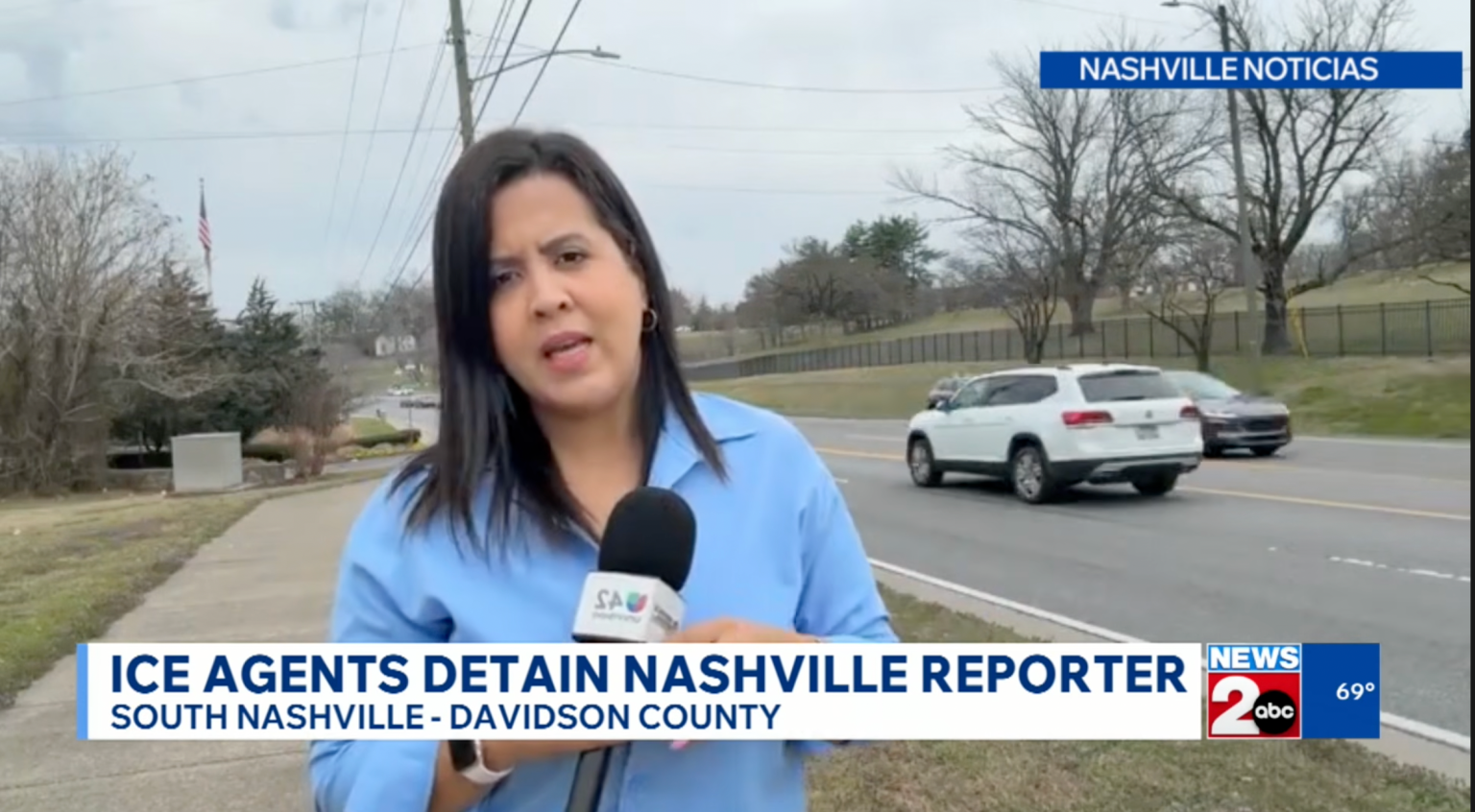 A female news reporter stands on a sidewalk holding a microphone, speaking to the camera. Cars drive by on the road behind her. The screen displays headlines about ICE agents detaining a Nashville reporter and news channel graphics.