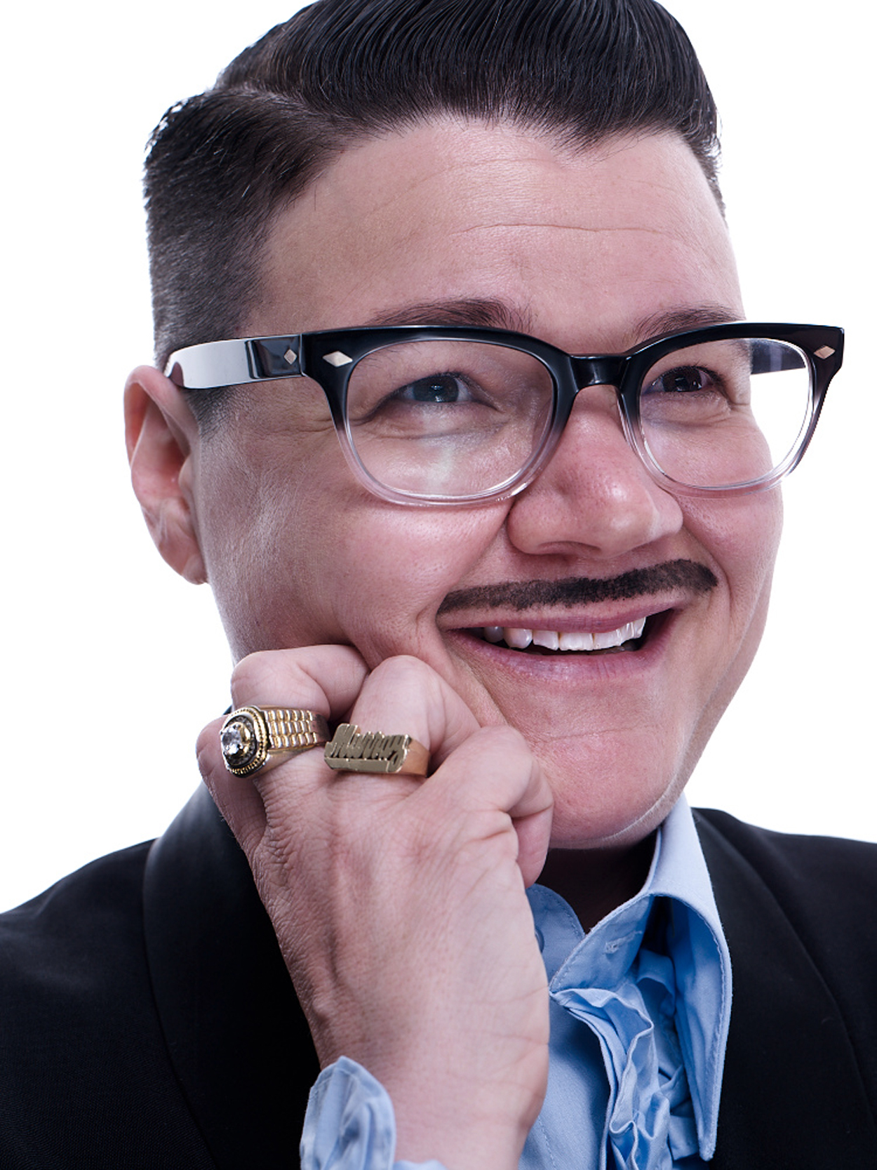 A person in a suit and glasses smiles widely, holding their chin with a hand wearing two large rings. The person has neatly styled dark hair and a thin mustache, and is wearing a light blue shirt with a cravat.