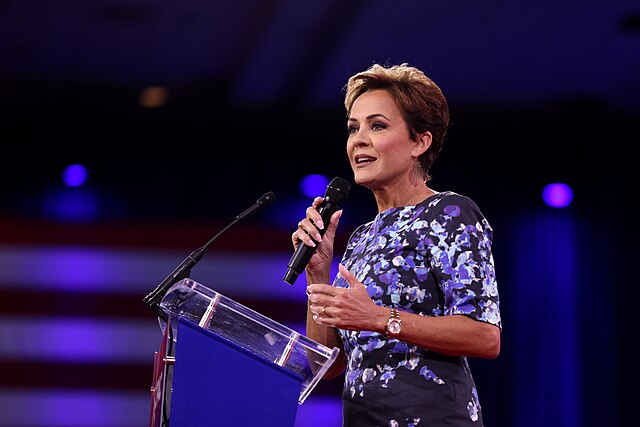 A woman in a blue floral dress speaks into a handheld microphone while standing at a clear podium, with blurred purple lighting and part of an American flag visible in the background.