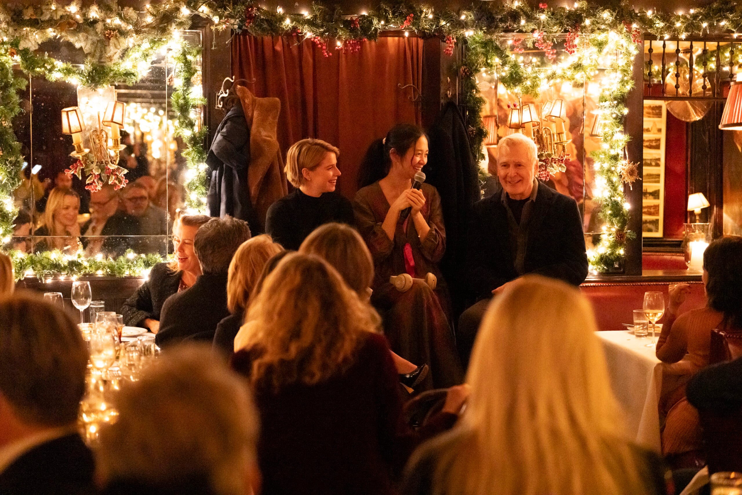 A warmly lit, festive room with holiday garlands and string lights hosts an event. Three people sit at the front, engaging with a seated audience, while others watch and enjoy the gathering.