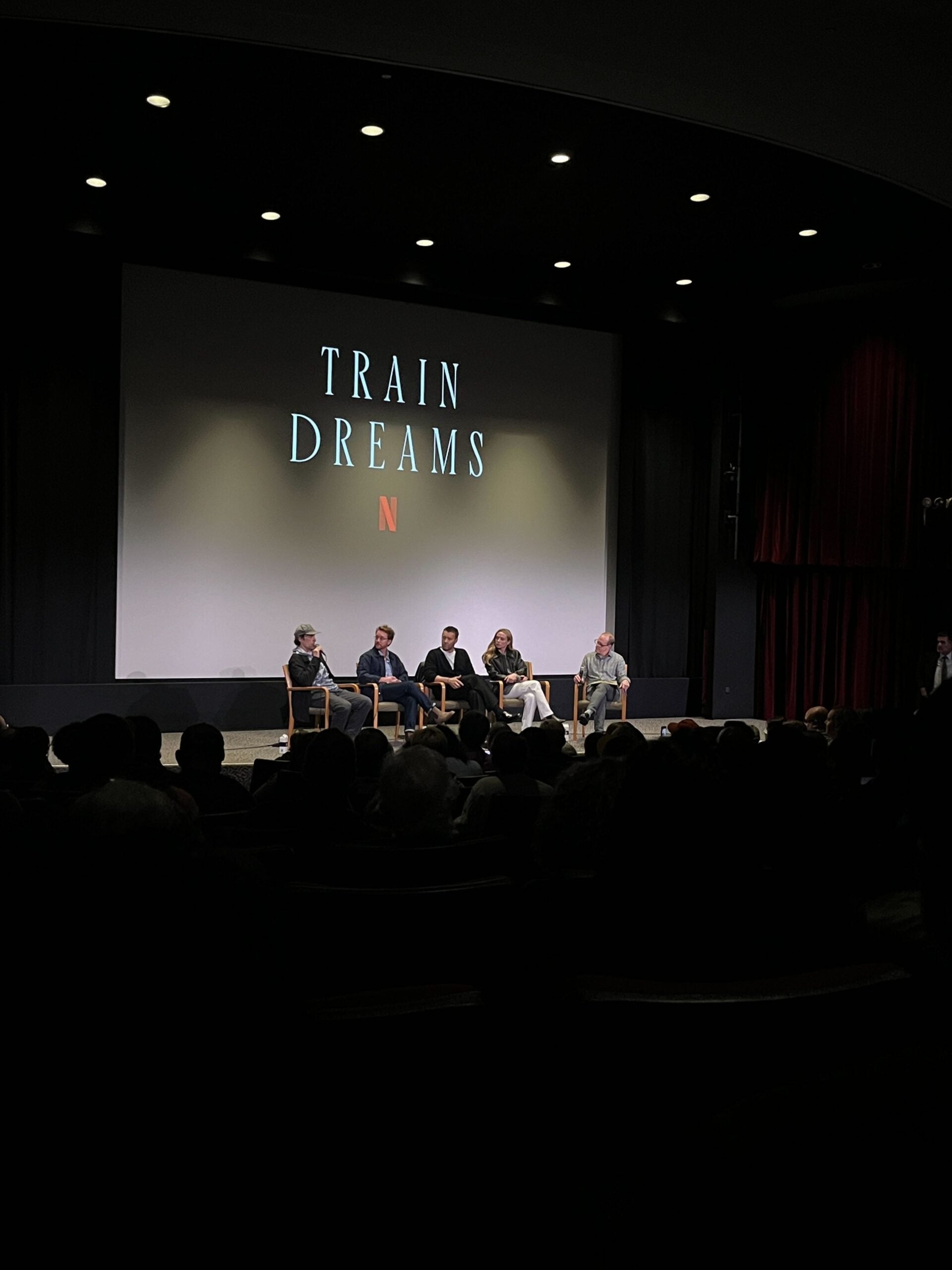Four people sit on stage in front of a screen displaying Train Dreams with a Netflix logo, as an audience watches in a darkened theater.