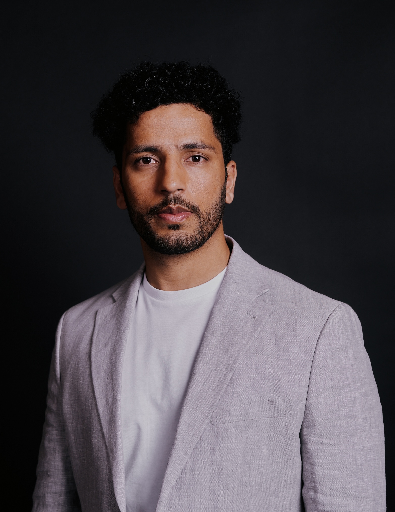 A man with short curly hair and a beard, wearing a light grey suit jacket over a white t-shirt, stands in front of a dark background, looking confidently at the camera.