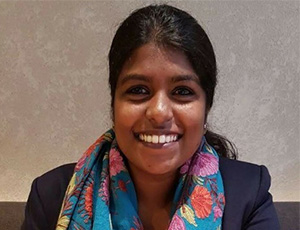 A woman with dark hair smiles at the camera. She is wearing a dark blazer and a colorful scarf with floral patterns, sitting against a light textured background.