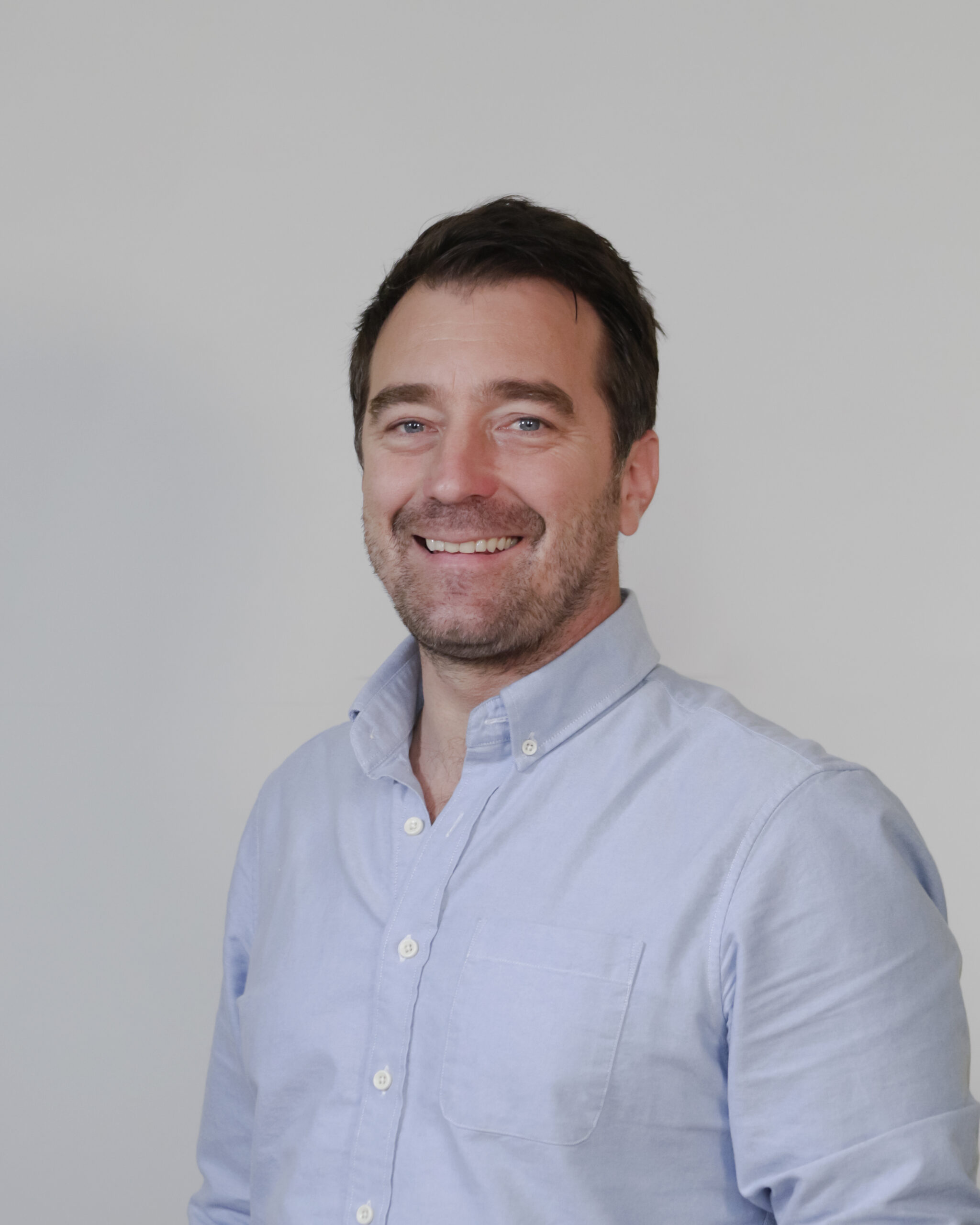 A man with short dark hair and a beard, wearing a light blue button-up shirt, smiling at the camera with a neutral gray background.