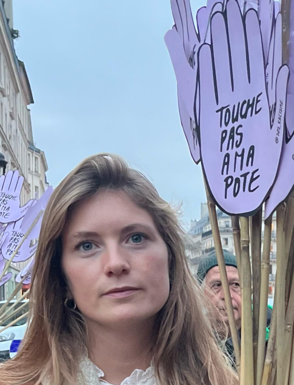 A woman stands outside holding purple hand-shaped signs that read Touche pas à ma pote during a protest or rally. Other people and buildings are visible in the background.