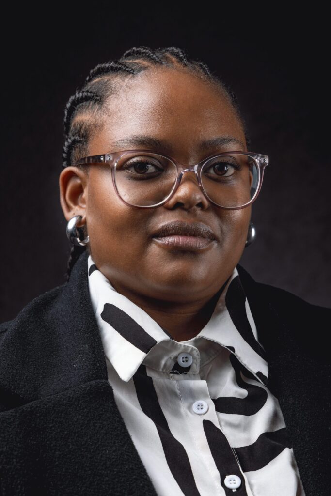 A person with braided hair, wearing glasses, hoop earrings, a black and white patterned shirt, and a dark jacket, poses against a dark background, looking confidently at the camera.